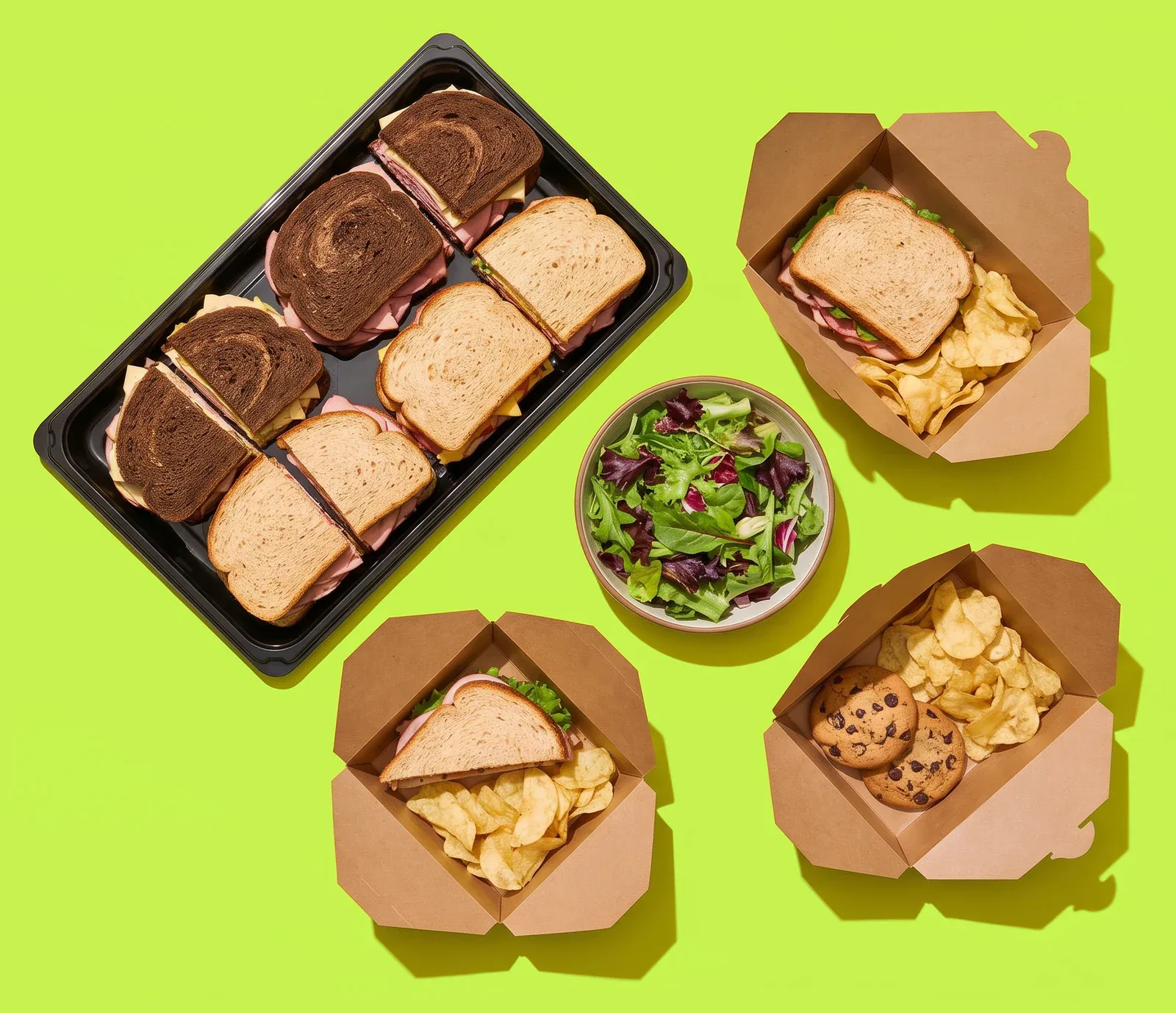 Deli sandwiches on a catering tray, a green salad, boxed lunches and cookies, shot from above