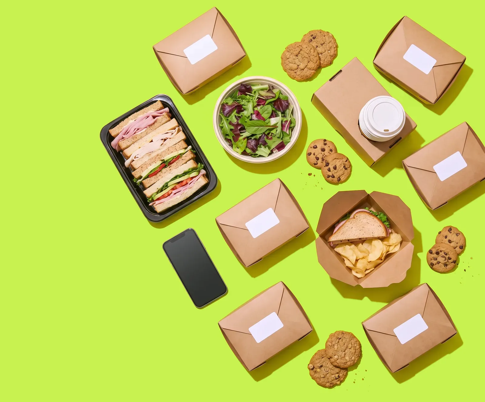 A catering order just delivered to an office: a tray of deli sandwiches, a green salad, labeled lunch boxes, coffee and cookies, with a phone lying among them